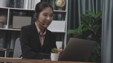 Kulaklık ve mikrofon takan iş kadını masasında çalışıyor ve dizüstü bilgisayar kullanıyor. Uzaktan kumandalı online toplantı için dizüstü bilgisayar kullanan hevesli kadın çalışanlar, ofiste iş videosu konferansı.