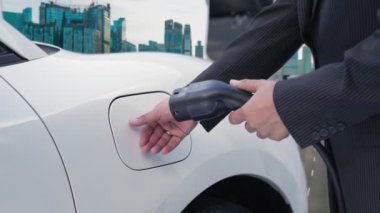 İlerici işadamı elektrikli aracını şarj kablosu ile şarj ediyor. Şarj istasyonundan şehrin arka planına bakıyor. Modern temiz enerji kampanyası konsepti için..