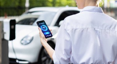 Suit-clad progressive businesswoman look at EV cars battery status from her phone while standing on a charging station with a power cable plug and a renewable energy-powered electric vehicle.