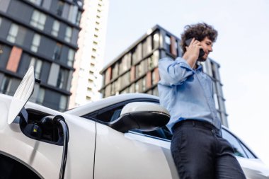 Progressive businessman talking on the phone, leaning on electric car recharging with public EV charging station, apartment condo residential building on the background as green city lifestyle.