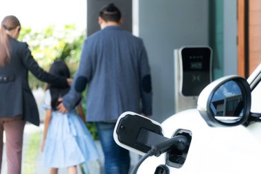 Focus closeup electric vehicle recharging battery from home electric charging station with blurred family in background. Renewable clean energy car for progressive eco awareness lifestyle concept.