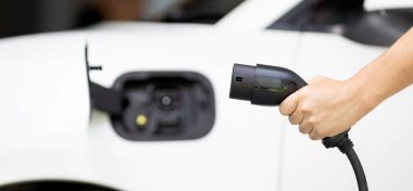 Focus hand holding EV charger plug with blurred background of progressive electric vehicle and socket parking in home garage with electric charging station powered by clean and sustainable energy.