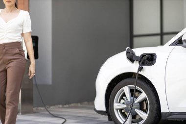 Focus image of electric vehicle recharging battery at home charging station with blurred woman walking in the background. Progressive concept of green energy technology applied in daily lifestyle.