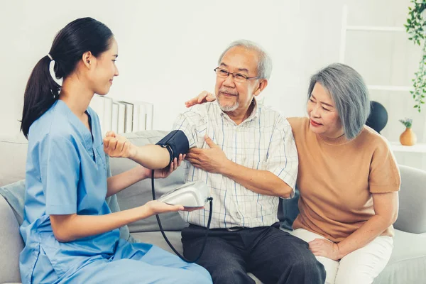 stock image An elderly man having a blood pressure check by his personal caregiver with his wife sitting next to him in their home.