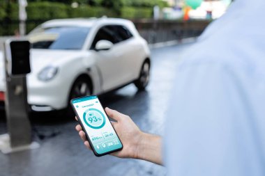 Closeup progressive businessman look at EV cars battery status application on smart phone screen at public parking charging station with power cable plug and renewable energy-powered electric vehicle