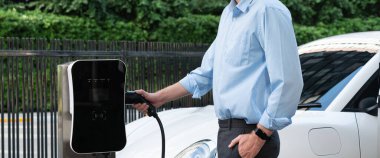 Closeup progressive man holding EV charger plug from public charging station for electric vehicle with background of residential building as concept eco-friendly sustainability energy car concept.