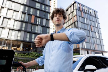 Businessman with smartwatch at modern charging station for electric vehicle with background of residential buildings as concept for progressive lifestyle of using eco-friendly as alternative energy.