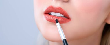 Closeup beautiful young woman with flawless healthy skin and natural makeup putting alluring fashion glossy red lipstick on her lip with lip brush in isolated background.