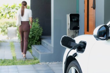 Focus EV charger plugged into EV car at home charging station with blurred background of progressive woman walking in background. Elective vehicle powered by clean energy for eco-friendly concept.