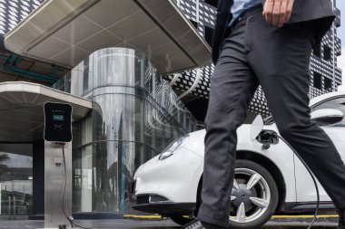 Closeup progressive businessman with electric car recharging at public charging station with background of city residential building. Eco friendly car powered by alternative clean energy.