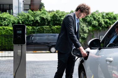 Modern şehir yerleşim bölgesindeki kamu şarj istasyonunda elektrikli araba şarj eden yenilikçi bir işadamı. Çevre dostu şarj edilebilir araba alternatif temiz enerjiyle çalışıyor..