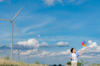 Progressive young asian boy playing with wind pinwheel toy in the wind turbine farm, green field over the hill. Green energy from renewable electric wind generator. Windmill in the countryside concept