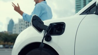Takım elbiseli, hırslı bir işadamı elektrik kablosu ve yenilenebilir enerji enerjili bir elektrikli araçla şarj istasyonunda dururken elektrikli aracına yaslanıyor..