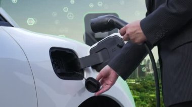 İlerleyen yeşil şehir ESG sembolü, elektrikli araçla birlikte. İş adamı EV arabalarını şarj etmek için şarj aletini şarj ediyor. Şehirdeki şarj istasyonunda ekolojik ve yeşil semboller var..