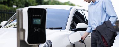 Closeup progressive man holding EV charger plug from public charging station for electric vehicle with background of residential building as concept eco-friendly sustainability energy car concept.