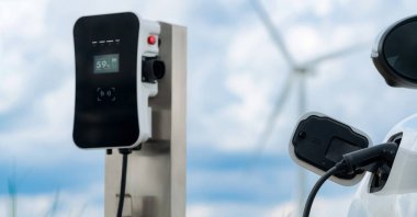 Progressive combination of wind turbine and EV car, future energy infrastructure. Electric vehicle being charged at charging station powered by renewable energy from wind turbine in the countryside.