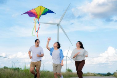 Progressive happy carefree family vacation concept. Young parents mother father and son run along and flying kite together on road with natural scenic on mountain and wind turbine background.