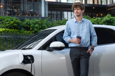 Progressive eco-friendly concept of parking EV car at public electric-powered charging station in city with blur background of businessman leaning on recharging-electric vehicle with coffee.
