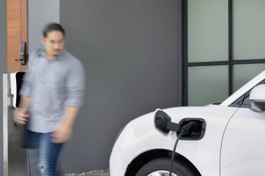 Focus electric car charging at home charging station with blurred progressive man walking in the background. Electric car using renewable clean for eco-friendly concept.