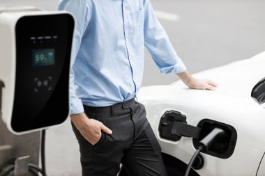 Closeup progressive suit-clad businessman with his electric vehicle recharge his car on public charging station in modern city with power cable plug and renewable energy-powered electric vehicle.