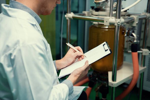 Closeup apothecary scientist using a clipboard and pen to record information from a CBD oil extractor and a scientific machine used to create medicinal cannabis products.