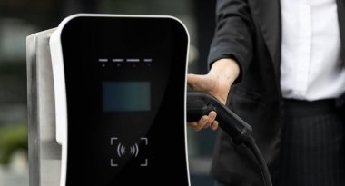 Focus EV charger plug and electric car at public charging station with blur progressive businesswoman holding charger and apartment condo building in background. Eco friendly electric vehicle concept.