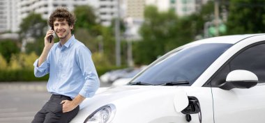 Progressive businessman talking on the phone, leaning on electric car recharging with public EV charging station, apartment condo residential building on the background as green city lifestyle.
