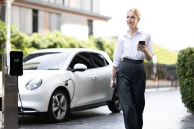 İş kadını tablet kullanıyor, halka açık otoparkta elektrikli aracını şarj ederken yürüyor. Teknolojinin ilerici yaşam tarzı ve EV otomobilinin ekolojik kaygısı