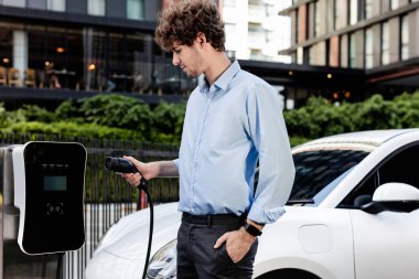 Progressive businessman insert charger plug from charging station to his electric vehicle with apartment condo building in background. Eco friendly rechargeable car powered by sustainable energy.