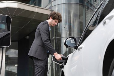 Below view closeup image of progressive black suit businessman recharge battery of his electric vehicle from public charging station. Renewable and alternative energy powered car concept.