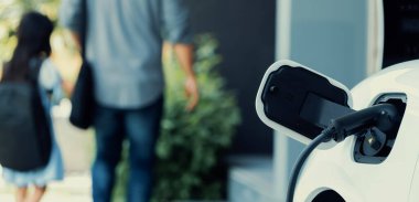 Arka planda bulanık baba ve kızının yürüdüğü şarj istasyonunda elektrikli araba şarj etmeye odaklan. Sağlıklı çevre yaşam tarzı konsepti için yenilikçi yeşil ve temiz enerji aracı.
