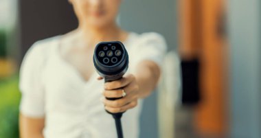 Focus hand holding and pointing EV charger plug for electric vehicle at camera with blur woman in background from home charging station as progressive idea of alternative energy for ecology concern.