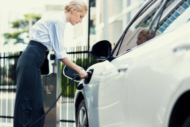 Progressive businesswoman insert charger plug from charging station to her electric vehicle with apartment condo building in background. Eco friendly rechargeable car powered by sustainable energy.