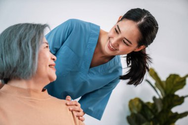 A caregiver rest her hands on the shoulders of a contented senior patient while she sitting on the sofa at home.