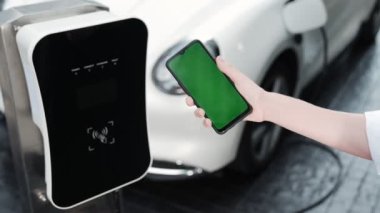 Taklit uzay için şarj istasyonuna bağlı elektrikli aracın yeşil ekran enerji durumunu gösteren bir model telefon. Temiz çevre için ilerici bir kavram. EV, yeşil yenilenebilir enerjiden güç alıyor.