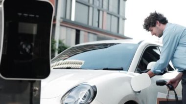 İlerici işadamı, şehir merkezinde dolaşmadan önce şarj cihazının fişini şarj istasyonundan elektrikli arabasına çekiyor. Çevre dostu şarj edilebilir araba, sürdürülebilir ve temiz enerjiyle çalışıyor..