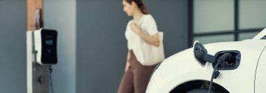 Focus image of electric vehicle recharging battery at home charging station with blurred woman walking in the background. Progressive concept of green energy technology applied in daily lifestyle.