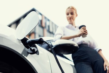 Şehir merkezinde dolaşmadan önce elektrikli arabası ve şarj istasyonu olan ilerici bir iş kadını. Çevre dostu şarj edilebilir EV araba sürdürülebilir ve temiz enerjiyle çalışıyor.