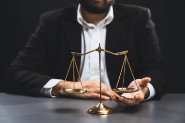 Siyah takım elbiseli bir avukat, altın bir denge, hukuki adalet ve dürüstlük sembolü, mahkemede dengeli ve etik karar verme mekanizması ile birlikte bürosunun masasında dikkatle oturuyor. denge