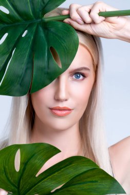 Closeup facial portrait personable woman with perfect smooth makeup holding green monstera leaves and cover her face as natural healthy skincare treatment. Tropical nature and beauty concept