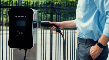 Closeup progressive man holding EV charger plug from public charging station for electric vehicle with background of residential building as concept eco-friendly sustainability energy car concept.