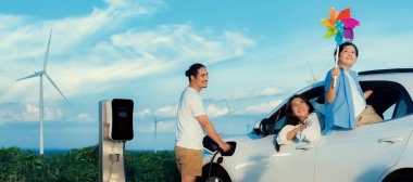 Concept of progressive happy family holding windmill toy and relax at wind farm with electric vehicle. Electric vehicle driven by clean renewable energy from wind turbine generator to charger station.