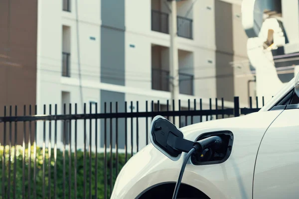 Closeup electric vehicle plugged in with EV charger at public charging station powered by renewable clean energy with residential building in background as progressive concept.