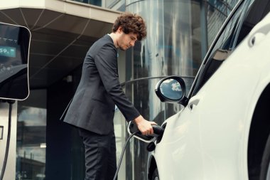 Below view closeup image of progressive black suit businessman recharge battery of his electric vehicle from public charging station. Renewable and alternative energy powered car concept.