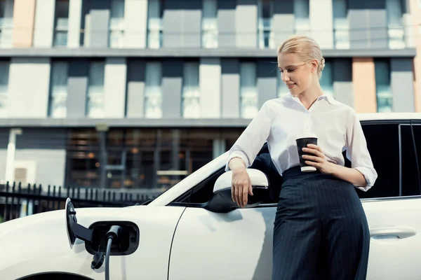 Businesswoman Drinking Coffee Leaning Electric Vehicle Recharging Public Charging Station — ストック写真