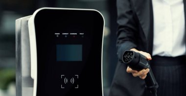 Focus EV charger plug and electric car at public charging station with blur progressive businesswoman holding charger and apartment condo building in background. Eco friendly electric vehicle concept.