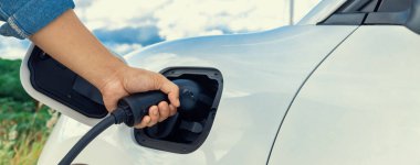 Progressive natural scenic with windmill generator where hand insert charging plug to electric vehicle from charging station with natural background. EV car powered by wind turbine electric generator.