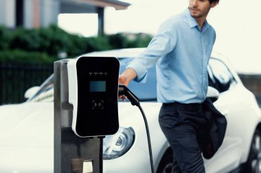 Progressive businessman insert charger plug from charging station to his electric vehicle with apartment condo building in background. Eco friendly rechargeable car powered by sustainable energy.