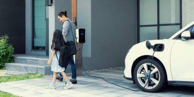 Progressive father and daughter plugs EV charger from home charging station to electric vehicle. Future eco-friendly car with EV cars powered by renewable source of clean energy.