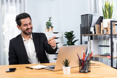 Siyah takım elbise giymiş heyecanlı ve mutlu bir iş adamı başarılı bir iş tanıtımı, enerji ve neşe kazandıktan sonra ofis masasında kutlama jesti olarak elini kaldırır. Coşkulu